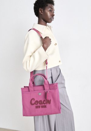 Femme portant une veste crème et un pantalon gris porte un sac cabas rose Coach New York avec bandoulière sur fond blanc.