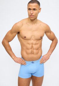 Μυώδης άντρας με κοντά μαλλιά, φορώντας ανοιχτό μπλε μποξεράκια Calvin Klein, στέκεται με τα χέρια στη μέση μπροστά σε μονόχρωμο φόντο.