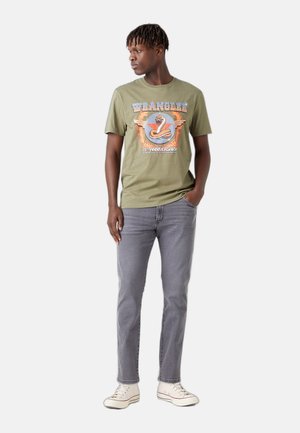 Giovane uomo che indossa una t-shirt grafica verde oliva Wrangler per il 75° anniversario, jeans grigi e sneakers bianche, in piedi con una mano in tasca.