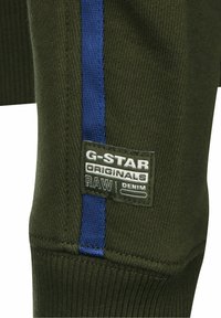 Tejido de punto verde oliva con una franja lateral azul y una etiqueta de G-Star Originals con letras blancas. Textura suave, diseño ajustado.