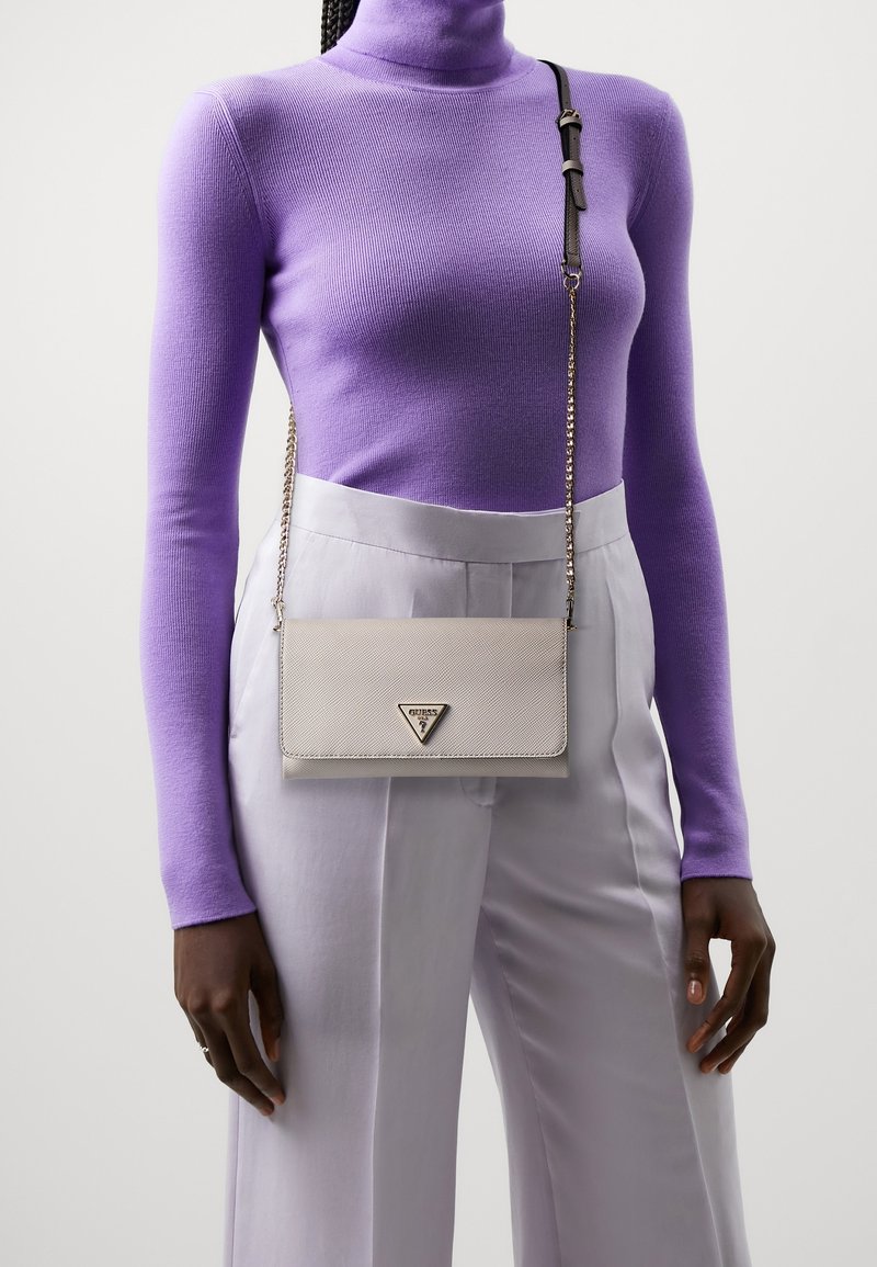 Lichtgrijze gestructureerde leren schoudertas met een driehoekig logo, zilveren kettingband en klep sluiting, gedragen over een paarse coltrui.