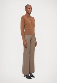 Pulover din tricot de culoare maronie, cu guler și detalii cu nasturi, asortat cu pantaloni largi cu talie înaltă din carouri maronii și pantofi cu toc negri.