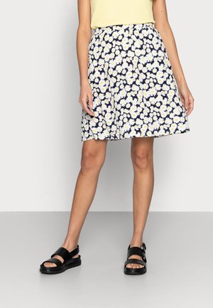 Vrouw die een marineblauwe rok draagt met een witte en gele madeliefjesprint, een lichtgele top en zwarte sandalen, staand op een grijze vloer tegen een witte muur.