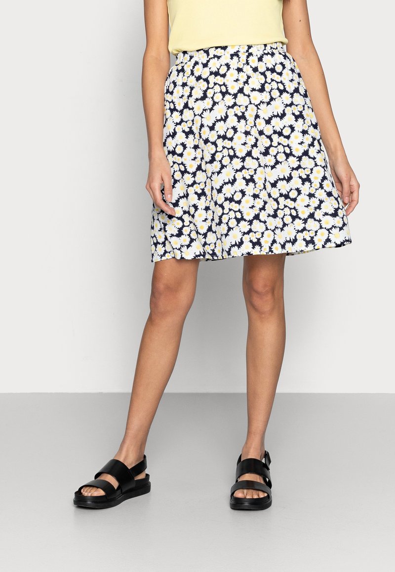 Femme portant une jupe bleu marine à imprimé marguerites blanches et jaunes, un haut jaune pâle et des sandales noires, debout sur un sol gris contre un mur blanc.
