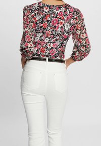 Blouse fleurie avec fond noir, fleurs roses et blanches, manches en voile transparent ; associée à un jean blanc ajusté et une ceinture noire ornée.