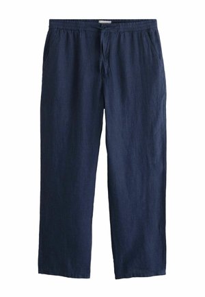 Férfi fekete rövid ujjú pulóverben, tengerészkék zsinóros nadrágban, fekete cipőben és napszemüvegben, kezében nagy tengerészkék táska fekete pántokkal.