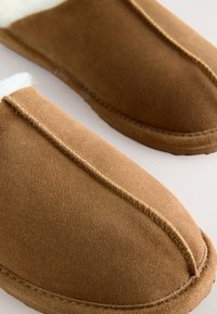 Brązowe zamszowe kapcie z białym sztucznym futerkiem, zaokrąglonym noskiem i przeszyciami wzdłuż szwów, o miękkiej fakturze.