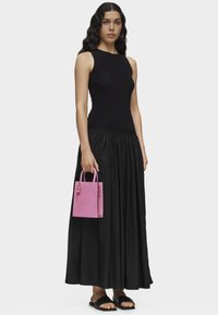 Top noir sans manches et jupe noire fluide avec un sac à main en cuir rose comportant un logo "TOUS" en relief et de petits pompons décoratifs.