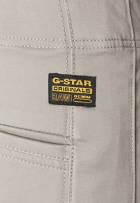 Bežs audums ar redzamām šuvēm, kuram ir melnas un dzeltenas krāsas marķējums ar uzrakstu "G-STAR ORIGINALS RAW DENIM." Smalka tekstūra.