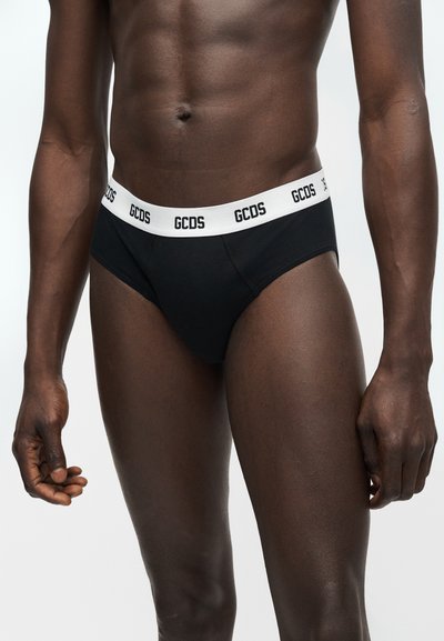 Torso masculin portant un slip noir avec une ceinture blanche affichant le logo "GCDS" répété sur un fond clair.