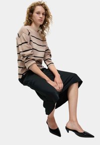 Beige randig tröja med svarta linjer, ihop med svarta cropped byxor. Modellen bär svarta spetsiga mule-skor. Lockigt hår är stylat löst.