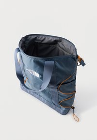 Μπλε τσάντα tote North Face με διπλές λαβές, κορυφή με ανοιχτό φερμουάρ, πλαϊνά λάστιχα και ευρύχωρο εσωτερικό σε λευκό φόντο.