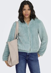 Kunstpelz-Jacke mit Reißverschluss in hellem Türkis, mit weicher Textur und gerippten Bündchen. Kombiniert mit einer beigefarbenen Tragetasche und blauen Jeans.