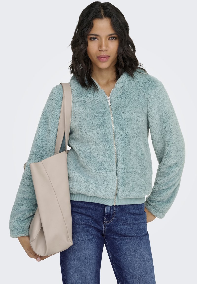 Kunstpelz-Jacke mit Reißverschluss in hellem Türkis, mit weicher Textur und gerippten Bündchen. Kombiniert mit einer beigefarbenen Tragetasche und blauen Jeans.