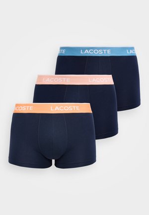 Tre boxer uomo Lacoste blu navy con fascia elastica in vita rispettivamente blu, rosa e arancione, ciascuno con il logo Lacoste in bianco.