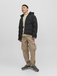 Giacca a vento nera con cappuccio, chiusura con zip e tasche laterali. Maglione beige sotto. Pantaloni cargo beige chiaro. Scarpe da ginnastica grigie.