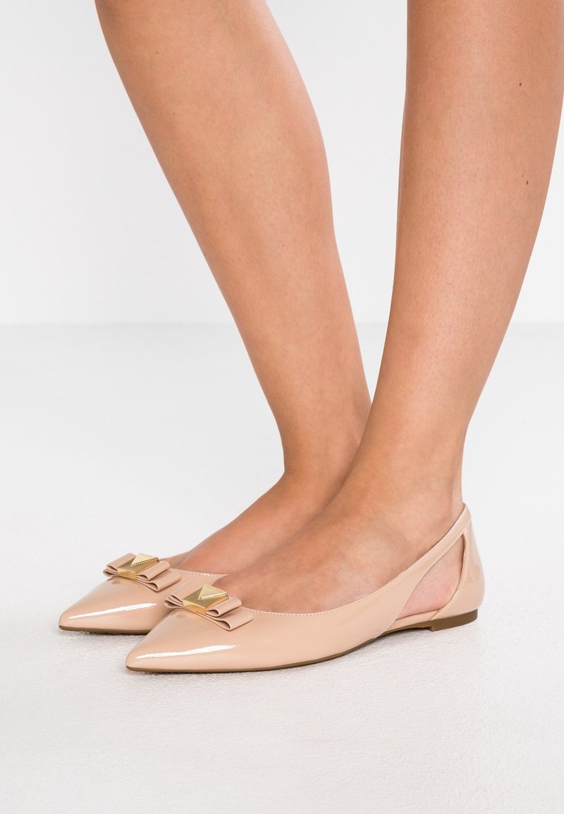 Chaussures plates beiges en cuir verni à bout pointu avec bride arrière, décorées de nœuds et de clous pyramides dorés, portées pieds nus.