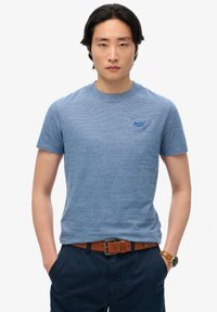 Maglietta a maniche corte a righe blu con scollatura a girocollo e un piccolo logo sul lato sinistro del petto, indossata con pantaloni blu scuro e una cintura marrone.