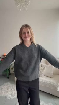 Šedý nadrozmerný sveter s V-výstrihom, dlhými rukávmi a jemnou textúrou, v kombinácii s čiernymi nohavicami. V pozadí je svetlý gauč.