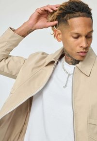 Jeune personne avec des dreadlocks portant une veste beige et une chemise blanche, ajustant ses cheveux d'une main, montrant des tatouages au cou et un collier de perles.