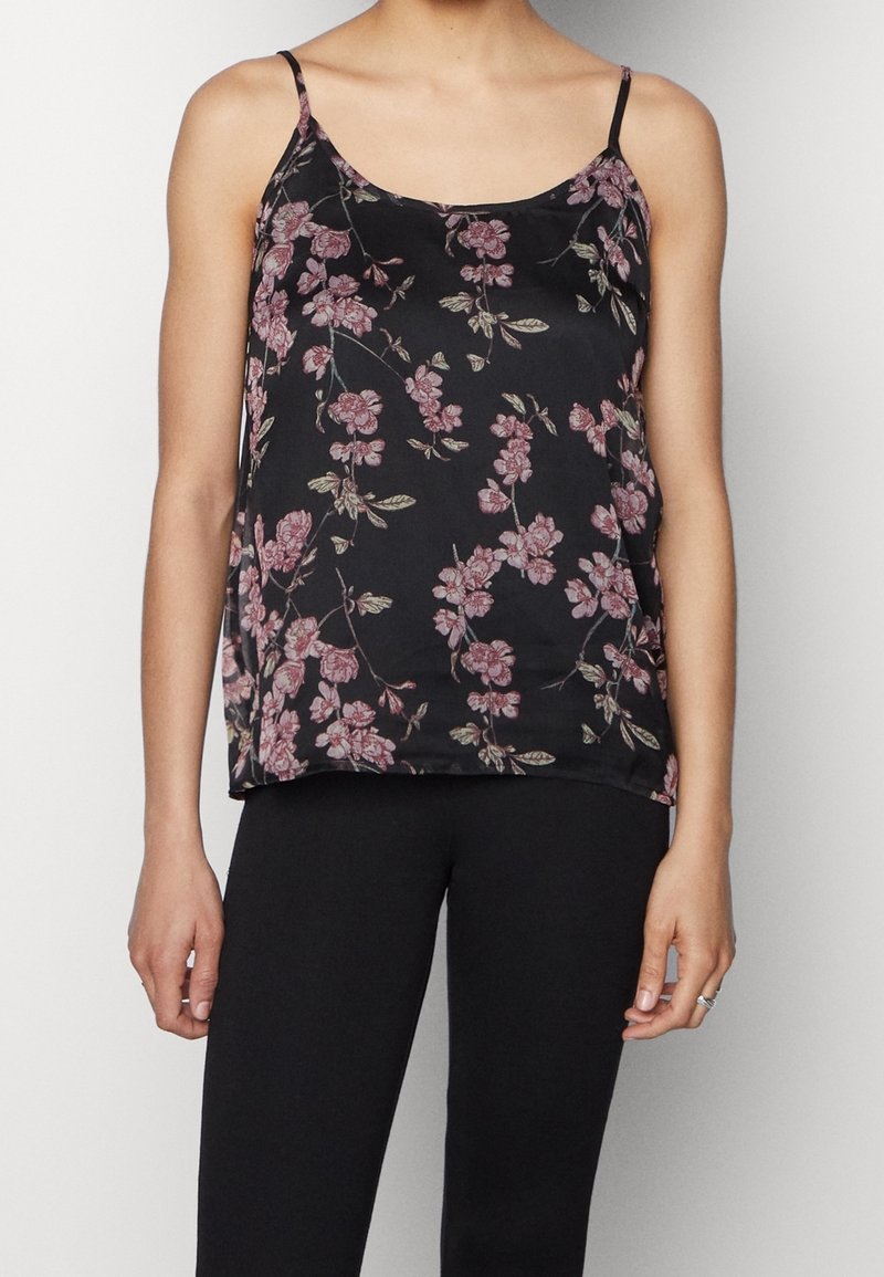 Femme portant un haut noir à bretelles fines orné de fleurs roses et un pantalon noir, debout devant un fond uni.