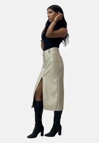 Falda beige hasta la rodilla con abertura lateral, cintura alta, pasadores para cinturón y bolsillos, combinada con botas negras de tacón alto hasta la rodilla. Color sólido, textura suave.