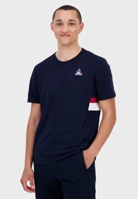 T-shirt en coton bleu marine avec col rond, présentant un petit logo sur la poitrine et des rayures horizontales rouges et blanches sur la manche.