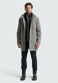 Grau melierter Wollmischmantel mit einer abnehmbaren schwarzen Innenjacke, Knöpfen und einem hohen Kragen. Getragen über dunklen Hosen und schwarzen Stiefeln.
