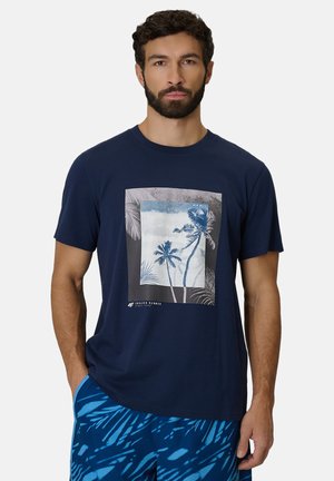 Mężczyzna z krótkimi kręconymi włosami i brodą, ubrany w granatowy t-shirt z nadrukiem palmy oraz niebieskie szorty w wzory, stoi z jedną ręką w kieszeni.