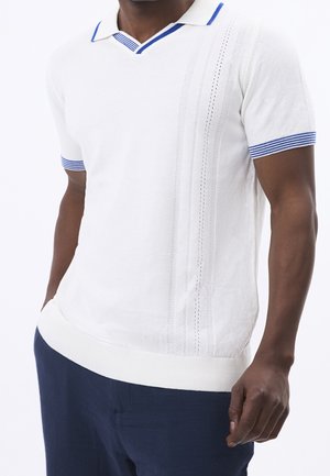 Mand iført hvid kortærmet strikket polo med blåstribet krave og manchetter, kombineret med mørkeblå bukser, hænder afslappet langs siden.