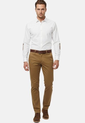 Hombre de pie mirando al frente, vestido con una camisa blanca con botones, cinturón marrón, pantalones beige y zapatos de ante marrón, sobre un fondo liso y claro.