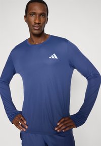 Αθλητικό τοπ με μακριά μανίκια σε σκούρο μπλε, κατασκευασμένο από λείο ύφασμα. Διαθέτει λευκό λογότυπο στο στήθος και εφαρμοστή σιλουέτα.