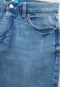 Gros plan sur un jean en denim bleu montrant la poche avant, la passants de ceinture et un bouton argenté avec des détails de couture visibles.