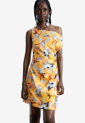 Femeie îmbrăcată într-o rochie fără mâneci, cu un umăr gol, cu un model floral galben, portocaliu și albastru și cercei mari albi în formă de floare.