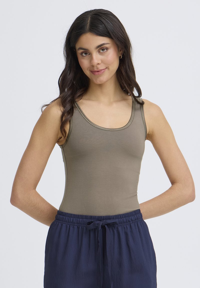 Jeune femme aux longs cheveux foncés portant un débardeur taupe et un pantalon à cordon bleu marine, se tenant les mains derrière le dos devant un fond uni.
