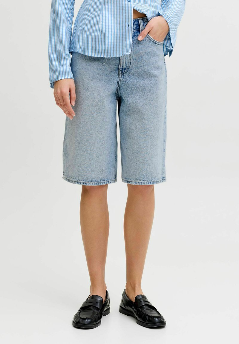 Inimene, kes kannab põlvepikkuseid helesiniseid teksapükse, sinist triibulist särki ja musti libisevaid mokasiine, seisab valgel taustal.