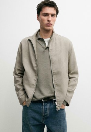 Giovane uomo con capelli corti e scuri che indossa una giacca beige con zip, polo verde oliva, jeans blu, mani in tasca, in piedi davanti a uno sfondo bianco.