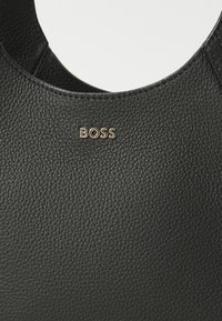 Sac à main en cuir noir avec une finition texturée ; présente un accent doré avec le logo "BOSS" près du haut. Design ouvert et courbé. Doublure intérieure lisse.