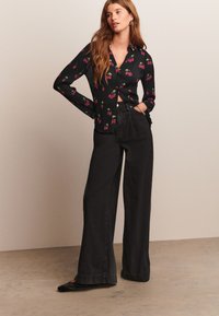 Chemise noire à fleurs avec motif cerises, manches longues, style boutonné, associée à un pantalon noir taille haute à jambes larges et des ballerines noires.