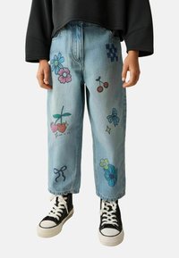 Lyseblå denimjeans med farverige blomster- og kirsebærgrafikker. Har en afslappet pasform og manchetter i bunden. Sammen med sorte høj-top sneakers.