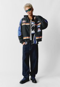 Crna jakna s plavim i narančastim naglasima, s logotipima. Nosena preko bijele majice, u kombinaciji s širokim trapericama i crnim čizmama.