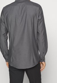 Chemise à manches longues grise à motif avec poignets à boutons, présentant un tissu texturé et un ourlet arrondi, vue de dos.