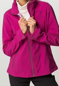 Persona con una chaqueta magenta de cremallera sobre un cuello de tortuga blanco, con las manos agarrando el cuello de la chaqueta cerca de la cremallera.