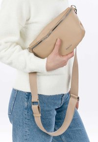 Beige kožená kabelka přes rameno s texturovaným povrchem, stříbrným zipem a nastavitelým látkovým popruhem, kterou drží člověk v bílém svetru.