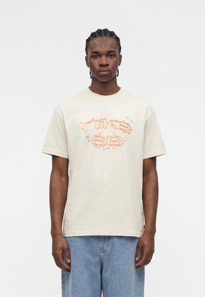 Giovane uomo con capelli intrecciati che indossa una t-shirt Nike SB color crema con stampa in stile graffiti arancione e jeans azzurri, in piedi davanti a uno sfondo bianco.
