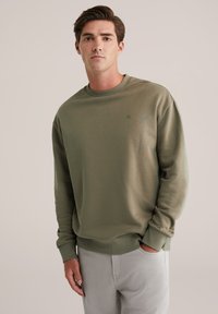 Olično zelena majica s kapuljačom iz mehkih tkanin z okroglim ovratnikom, rebrastimi manšetami in majhnim logotipom na prsih. V kombinaciji s svetlo sivo hlačami.