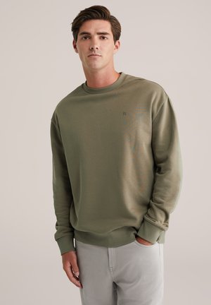 Sweatshirt vert olive en tissu doux avec un col rond, des poignets côtelés et un petit logo sur la poitrine. Assorti avec un pantalon gris clair.
