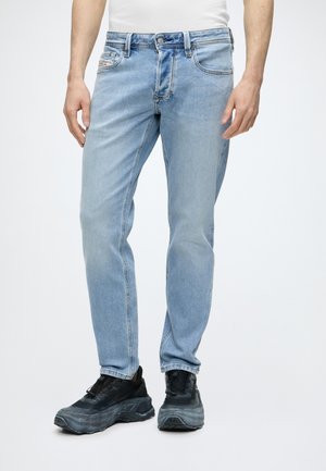 Homme portant un jean droit bleu clair et des baskets noires épaisses, debout devant un fond blanc uni.