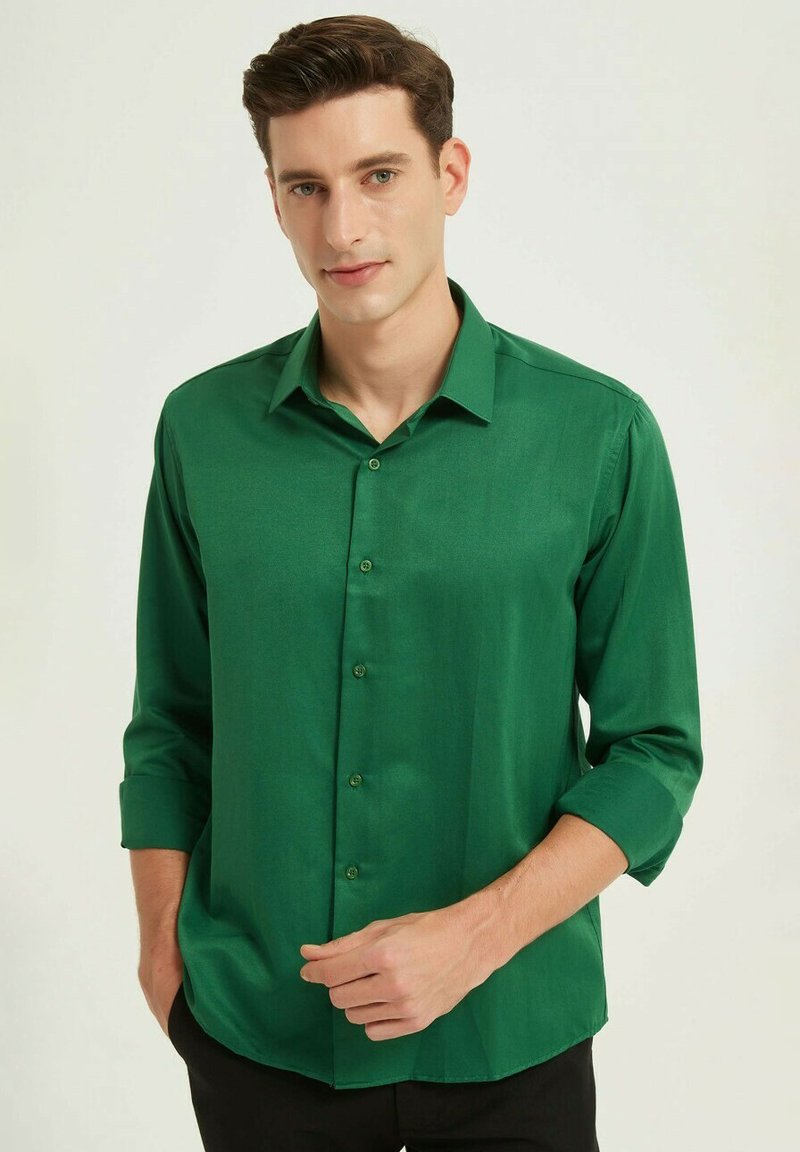 Chemise vert à boutons avec une texture lisse, manches longues retroussées aux coudes et col classique. Coupe décontractée, associée à un pantalon foncé.