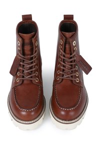 Botas de cuero marrón hasta el tobillo con ojales de metal, cordones, suelas de goma blanca y costuras decorativas. Incluye una etiqueta de marca en el lateral.